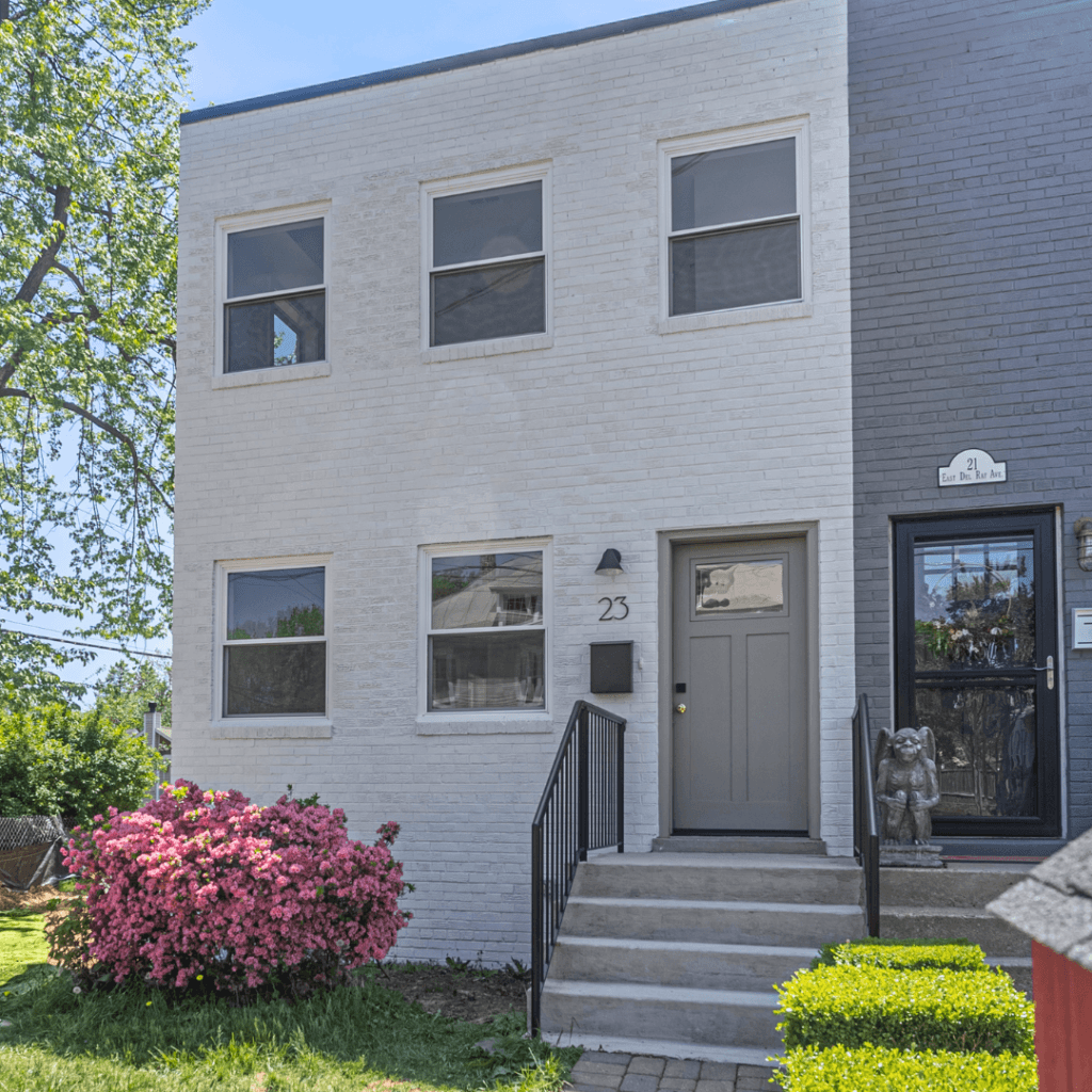 A photo showcasing the exterior of a renovated brick home. The facade has been updated with a fresh coat of light gray paint, and the entryway features a modern gray door with black hardware and a matching black light fixture. The front yard is neatly landscaped with green grass and a blooming pink azalea bush near the entrance. The walkway is paved with bricks, leading up to the front steps with black railings, giving the home a clean and inviting appearance