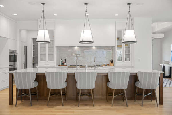 The image shows a modern kitchen with a large island. The island has a wooden base and a light-colored countertop, with five upholstered bar stools featuring a light fabric with vertical stripes. Above the island, there are three large pendant lights with white shades. In the background, there's a white kitchen with integrated appliances, a range hood over a stove, and open shelving with glass-front cabinets. The flooring is light-colored hardwood.